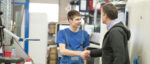 Schüler in blauer Latzhose schüttelt in Werkstatt einem älteren Mann die Hand.
